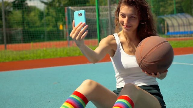 Athletic girl doing selfie with ball on smartphone while sitting on sports field
