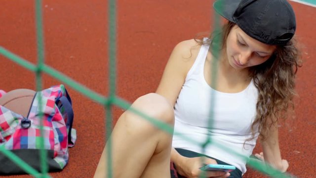 Girl sitting behing net on the sports field and using smartphone
