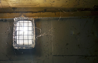  Shining old light covered in metal grill and cobwebs against dirty, peeling painted wall.