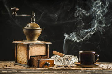 A cup of coffee with an old grinder and smoking a cigar in the background
   


