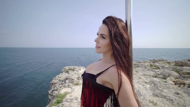 Young woman pole dancer in red bodysuit with portable stand alone fitness dance pole on a rock above the sea. Tarhankut cape, steadicam shot 4k