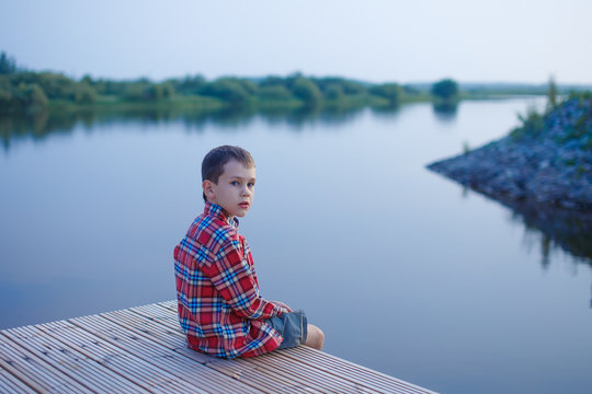 boy looks cautiously sitting near the water. child sitting on a wooden pier on the background of the river. quiet evening. boy startled looks back