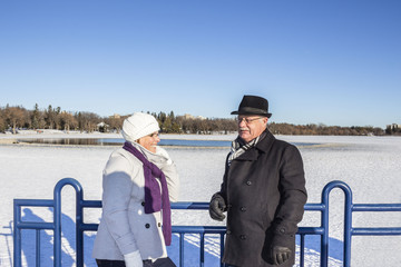 horizontal image of a man and woman wearing winter coats and hats leaning against a blue steel railing chatting on a sunny cold winter day.