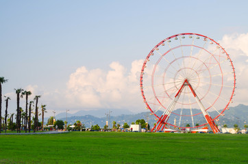 ferris wheel on green field, mountains, palms and blue sky with light clouds in Batumi, Georgia