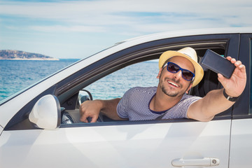 Portrait of young tourist guy taking selfie with smart phone from driver’s seat. Ocean in the background. Travel and transportation concepts.  