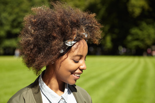 Profile of pretty dark-skinned student girl with curly stylish hair, wearing do-rag, having morning walk along green lawn in urban forest, closing her eyes because of bright sunshine, smiling happily