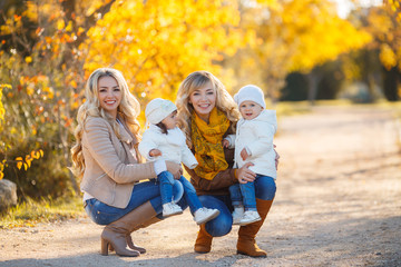 Two young mothers,a blonde,dressed in blue jeans and leather jackets,walk in the Park day on the background of beautiful yellow autumn nature with young daughters,dressed in white jackets