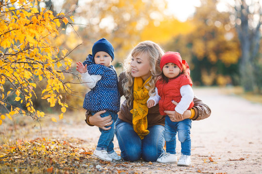 Young mother with long curly blond hair and blue eyes, dressed in blue jeans walking in yellow autumn Park with two young daughters,dressed in blue jeans,red and blue jackets