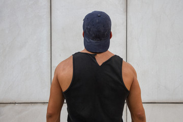 Young, caucasian, strong and handsome man wearing a black tank top and a cap. He is looking at the white wall on the street and is giving back. 