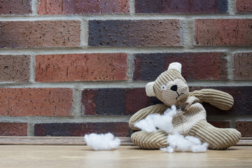 A sad, abandoned teddy bear with stuffing and filling falling from a hole in his ripped and torn tummy and sitting alone against a brick wall.