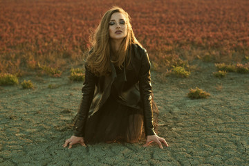 Young glam model with long hair in black veiling dress and stylish leather jacket sitting on the ground with closed eyes and getting Earth's energy at the sunset. Fashion and make up concept.