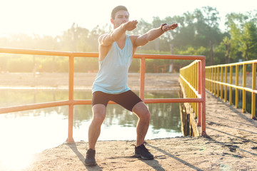 Fototapeta premium Fit man warming up doing squats stretching arms forward outdoors.