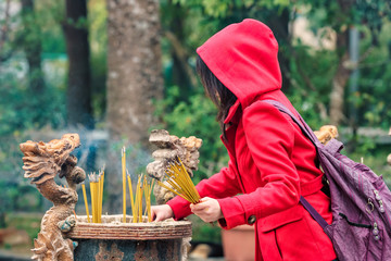Hooded figure of a young woman placing burning Buddhist prayer sticks into the urn at Po Lin Monastery on Lantau Island in Hong Kong