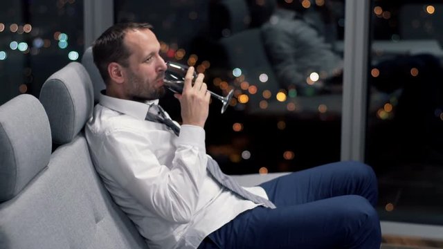Young businessman drinking red wine sitting on sofa at home at night
