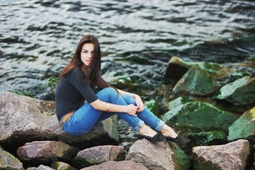 Beautiful young long-haired girl in jeans and a dark blouse posing on the rocks  the sea shore into the waves of an emerald color.