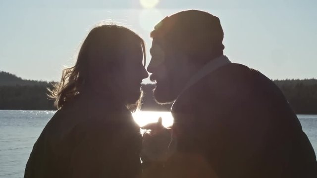 Backlit of young loving couple sitting near lake kissing and declaring love for each other 