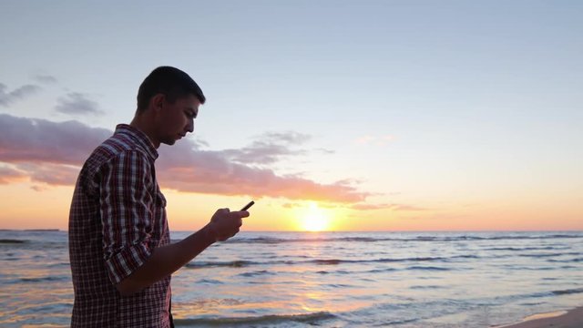Young lonely man walking along the seashore at sunset, typing a message on the phone