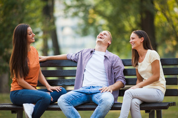 Three friends are sitting on bench in park and talking.