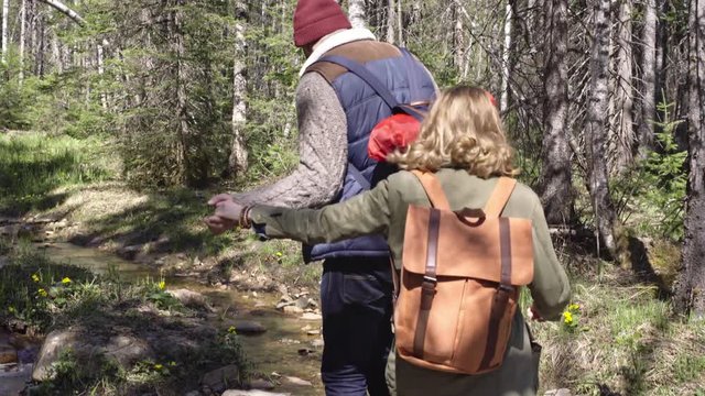 Handheld tracking shot of cheerful young couple with backpacks walking in forest and jumping over little brook