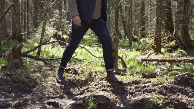 Handheld tracking with tilt down of bearded man walking around forest and stepping over brook 