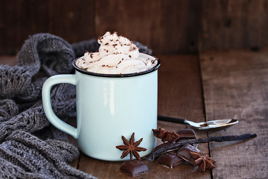 Cup of hot cocoa or coffee for Christmas with whipped cream, shaved chocolate, vanilla pod, spices and gray scarf against a rustic background. Shallow depth of field with selective focus on drink. 