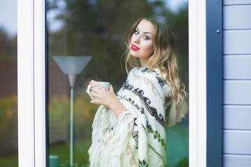 Fototapeta premium Young beautiful blonde woman with cup of coffee standing by the window. Garden reflection on the glass. 