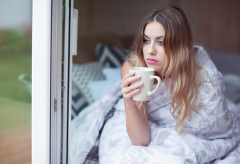 Young beautiful blonde woman with cup of coffee sitting home on the bed covered with duvet by the...