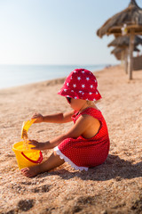Baby girl in red dress and hat playing toys on a beach