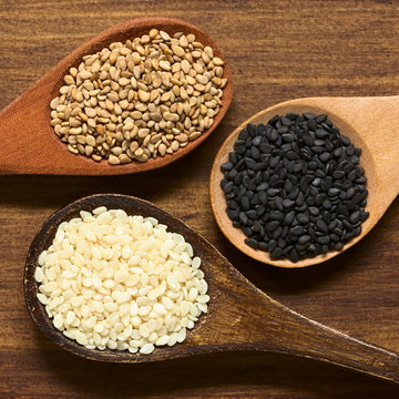 Black, white and roasted sesame seeds on small wooden spoons, photographed overhead on wood with natural light (Selective Focus, Focus on the seeds on the top)