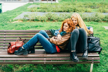 Two young beautiful caucasian redhead and blonde women friends sitting on a bench in a city park, ...