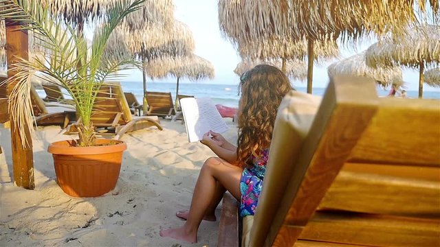 Cute little girl reading book among straw parasols and wooden seabeds on the beach