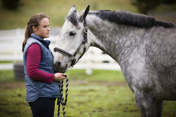 Young woman standing with her grey horse in a paddock.