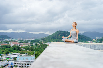 Fototapeta na wymiar Yoga on rooftop. Happy young woman in lotus pose on roof with city and mountains view.