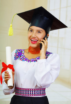 Young female student wearing traditional blouse with graduation hat, holding formal paper diploma roll while talking on mobile phone, smiling to camera