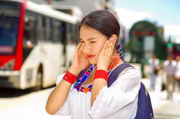 Pretty young woman wearing traditional andean blouse and blue backpack, waiting for bus at outdoors station platform, covering ears traffic noise, unpleasant facial expression