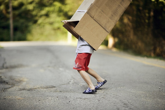 A small boy struggles to remove the box off his body while walking on the road.