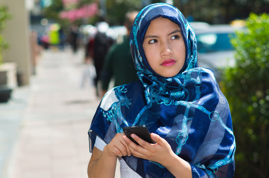 Beautiful young muslim woman wearing blue colored hijab, using mobile phone in outdoors urban background
