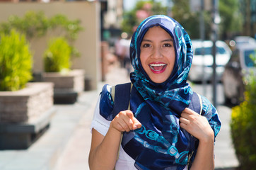 Beautiful young muslim woman wearing blue colored hijab, pointing finger smiling, outdoors urban...