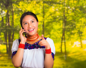 Beautiful young woman wearing traditional andean blouse with necklace, standing posing for camera, holding mobile phone talking, green forest background