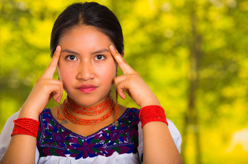 Headshot beautiful young woman wearing traditional andean blouse with red necklace, posing for camera touching face using hands while smiling happily, green forest background
