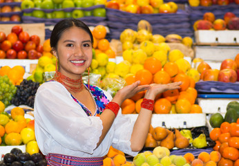 Beautiful young hispanic woman wearing andean traditional blouse posing holding hands out for camera inside fruit market, colorful healthy food selection in background