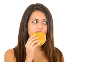 Portrait beautiful young woman posing for camera eating hamburger while making guilty facial expression, white studio background