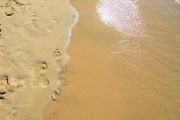 Fototapeta na wymiar Footprints on a sandy beach being washed away by the sea with the sun reflecting in the waves