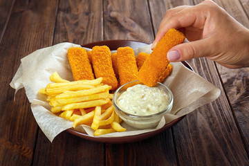 Woman holding tasty fish nugget and plate with fries and sauce on table