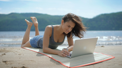 Fototapeta premium A beautiful sexy young smiling lady is laying on her tommy at a beach using laptop