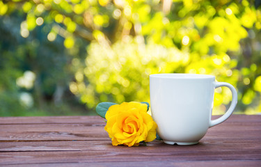 Obraz na płótnie Canvas White cup of tea and fragrant yellow rose on a wooden table. Romantic concept. Natural green background. Green blur. Copy space. 
