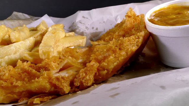 Dolly shot of a very British takeaway meal of fish and chips, with a hand dipping into the optional curry sauce.