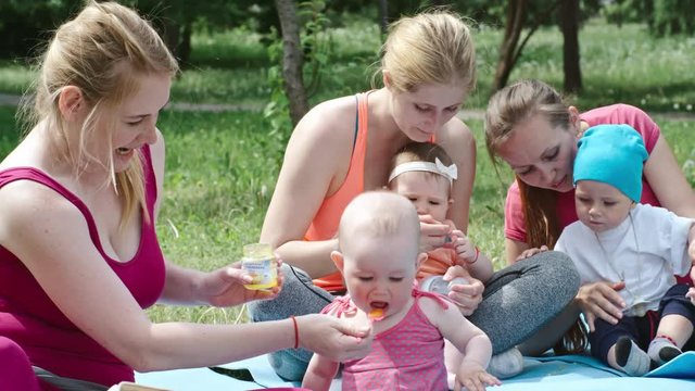 Four young women having picnic with their babies on green lawn at summer day: one woman feeding child with baby food from spoon, another kids eating cookies