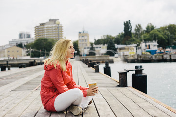 young blonde woman in red jacket sitting on the dock and holding cup of coffee