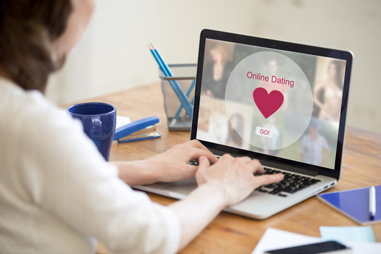 Young beautiful single woman browsing online dating website on a laptop display. Attractive woman model sitting with notebook, searching for new love online. Back view over the shoulder